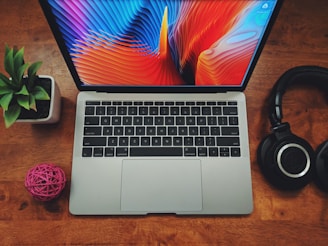 A neatly arranged setup featuring a laptop on a wooden surface. The laptop screen displays a vibrant, abstract multicolored pattern. To the left is a small green potted plant and a pink decorative ball. On the right, a pair of black headphones rests on the table.