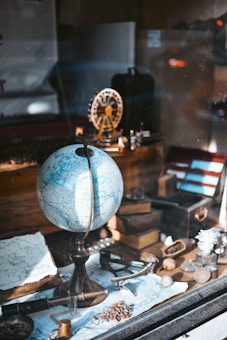 A vintage-themed setup featuring a globe prominently placed at the center, surrounded by various antique items including a map, an old book, a magnifying glass, and a small model of a Ferris wheel. The arrangement is set in a warm light, possibly a display window, conveying a sense of exploration and nostalgia.