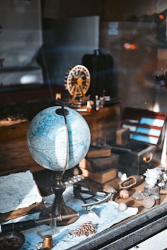 A vintage-themed setup featuring a globe prominently placed at the center, surrounded by various antique items including a map, an old book, a magnifying glass, and a small model of a Ferris wheel. The arrangement is set in a warm light, possibly a display window, conveying a sense of exploration and nostalgia.
