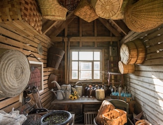 A rustic wooden shed with slatted walls houses an assortment of woven baskets hanging from the ceiling. Various gardening tools and containers, such as watering cans, are arranged on a table beneath a large window. A collection of brown glass bottles and a small arrangement of sunflowers add to the vintage decor.