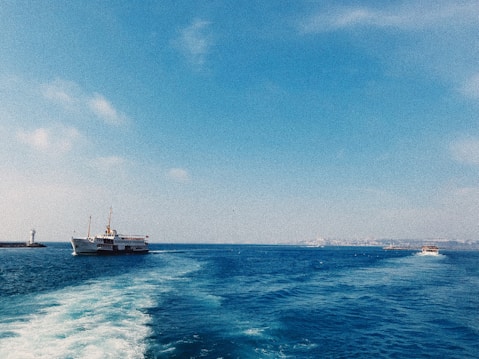 A large passenger boat is sailing across a calm blue sea, leaving a white trail in its wake. In the distance, another boat is heading toward the horizon. A lighthouse stands on a stone jetty on the left side, under a clear blue sky with a few scattered clouds.