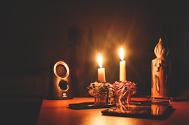 A warm, dimly lit setting with two lit candles casting a soft glow. A small speaker is positioned to the left, while decorative items, including a floral arrangement and a sculpted candle, are arranged on a wooden table.