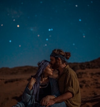 man and woman sitting on desert sand under blue sky during nighttime