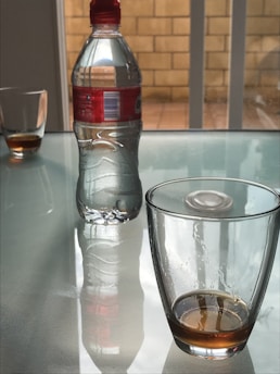 A clear glass with a small amount of brown liquid sits on a glass table, accompanied by a plastic water bottle with a red cap and label. Reflections of the bottle and glasses are visible on the table surface. In the background, there is a brick wall and glass sliding doors, creating a neutral environment.