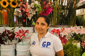 A person wearing a white polo shirt stands in front of a variety of colorful flowers arranged in white buckets. Sunflowers, lilies, and other blossoms create a vibrant and lush backdrop.