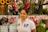 A person wearing a white polo shirt stands in front of a variety of colorful flowers arranged in white buckets. Sunflowers, lilies, and other blossoms create a vibrant and lush backdrop.