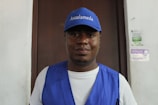 A man wearing a blue cap and vest is standing in front of a wooden door. The cap has 'Asoalameda' printed on it. The background includes posters on a white wall.
