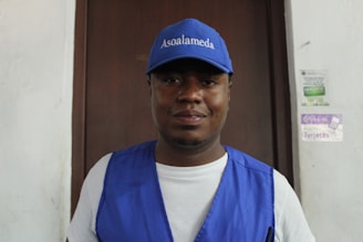 A man wearing a blue cap and vest is standing in front of a wooden door. The cap has 'Asoalameda' printed on it. The background includes posters on a white wall.