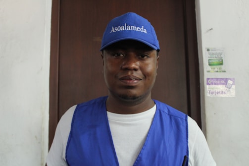 A man wearing a blue cap and vest is standing in front of a wooden door. The cap has 'Asoalameda' printed on it. The background includes posters on a white wall.