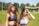 Two young women are standing on an athletic track. They are wearing sports bras, one with curly hair and the other with straight hair. The background shows a grassy field and trees under a clear blue sky.