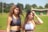 Two young women are standing on an athletic track. They are wearing sports bras, one with curly hair and the other with straight hair. The background shows a grassy field and trees under a clear blue sky.