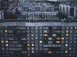 A large residential complex featuring multiple multi-story apartment buildings with numerous windows, some of which are illuminated. The buildings have a grey and slightly aged facade, and the scene is set during twilight or evening, as evidenced by the soft, dim lighting.