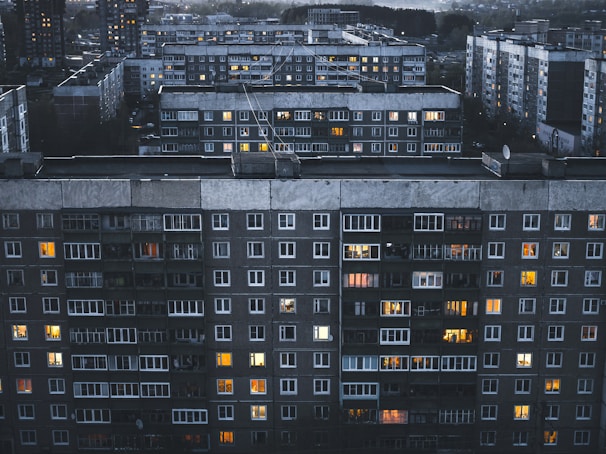 A large residential complex featuring multiple multi-story apartment buildings with numerous windows, some of which are illuminated. The buildings have a grey and slightly aged facade, and the scene is set during twilight or evening, as evidenced by the soft, dim lighting.