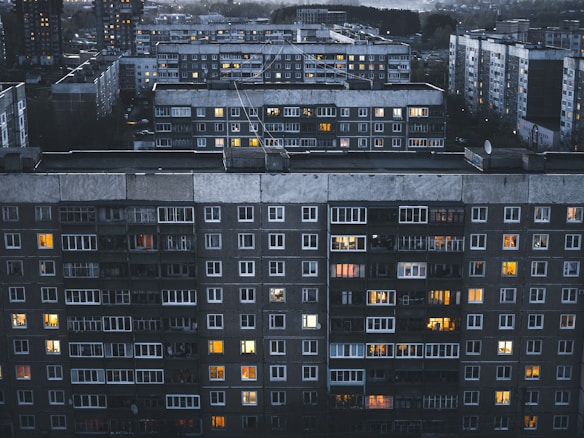 A large residential complex featuring multiple multi-story apartment buildings with numerous windows, some of which are illuminated. The buildings have a grey and slightly aged facade, and the scene is set during twilight or evening, as evidenced by the soft, dim lighting.