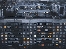 A large residential complex featuring multiple multi-story apartment buildings with numerous windows, some of which are illuminated. The buildings have a grey and slightly aged facade, and the scene is set during twilight or evening, as evidenced by the soft, dim lighting.