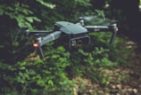 A drone with a sleek, modern design hovers in the air against a backdrop of lush green foliage. It has four rotors, each emitting a soft blur from the rapid movement. Two small lights on the front arms emit a soft red glow, possibly for navigation or signaling.