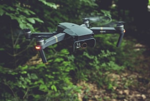 A drone with a sleek, modern design hovers in the air against a backdrop of lush green foliage. It has four rotors, each emitting a soft blur from the rapid movement. Two small lights on the front arms emit a soft red glow, possibly for navigation or signaling.