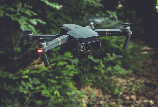 A drone with a sleek, modern design hovers in the air against a backdrop of lush green foliage. It has four rotors, each emitting a soft blur from the rapid movement. Two small lights on the front arms emit a soft red glow, possibly for navigation or signaling.