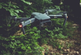 A drone with a sleek, modern design hovers in the air against a backdrop of lush green foliage. It has four rotors, each emitting a soft blur from the rapid movement. Two small lights on the front arms emit a soft red glow, possibly for navigation or signaling.