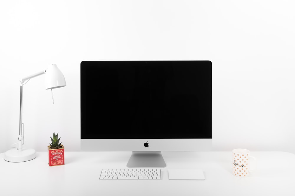 silberner iMac, Apple Wireless Keyboard und Apple Magic TrackPad in der Nähe der Tischlampe