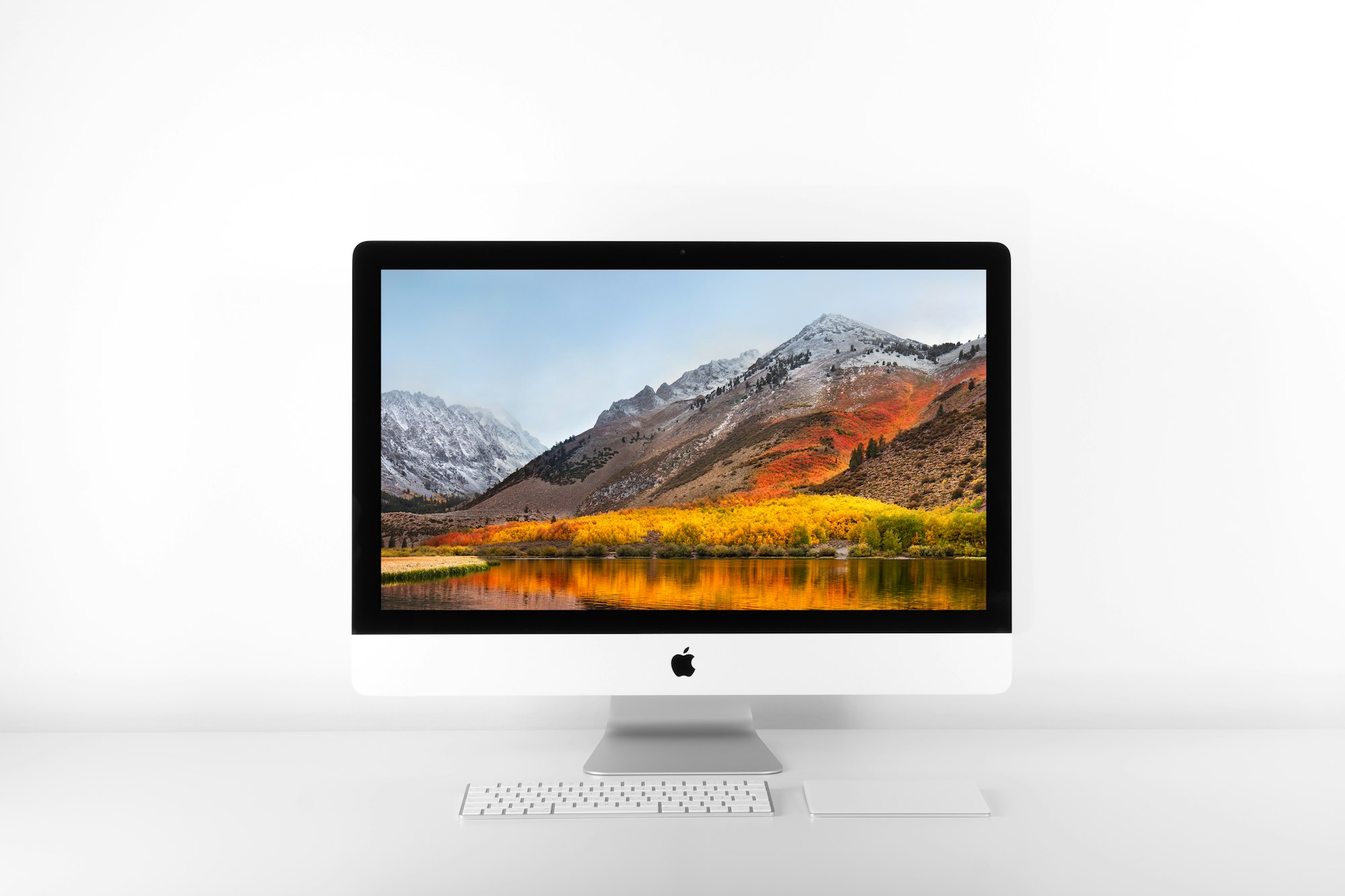 A dekstop iMac with a keyboard and trackpack on the desk in front. A lake and mountain picture is on the screen.