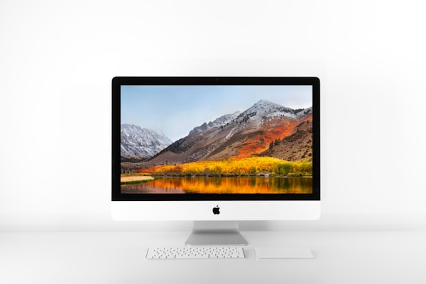 A dekstop iMac with a keyboard and trackpack on the desk in front. A lake and mountain picture is on the screen.