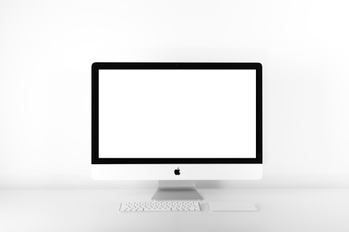 A minimalist setup featuring a desktop computer with a large, blank screen. The computer is an all-in-one model with a sleek, white design and is accompanied by a compact keyboard and a trackpad. The setup is positioned against a stark white background, emphasizing simplicity and modern aesthetic.