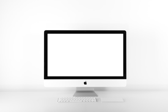 A minimalist setup featuring a desktop computer with a large, blank screen. The computer is an all-in-one model with a sleek, white design and is accompanied by a compact keyboard and a trackpad. The setup is positioned against a stark white background, emphasizing simplicity and modern aesthetic.