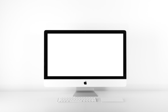 A minimalist setup featuring a desktop computer with a large, blank screen. The computer is an all-in-one model with a sleek, white design and is accompanied by a compact keyboard and a trackpad. The setup is positioned against a stark white background, emphasizing simplicity and modern aesthetic.