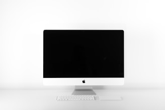 A sleek, modern desktop computer with a large, black screen and a minimalistic white frame, positioned on a white surface. In front of the screen is a compact white keyboard and a matching trackpad.
