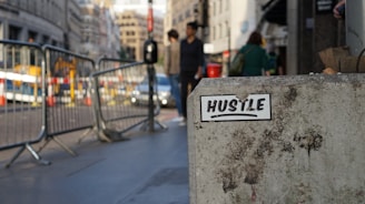 A concrete block with a sticker reading 'HUSTLE' in bold lettering is in the foreground, slightly weathered and positioned along a city street. Metal barriers line the sidewalk, and in the background, there are people walking and cars driving, with buildings densely packed along the road.