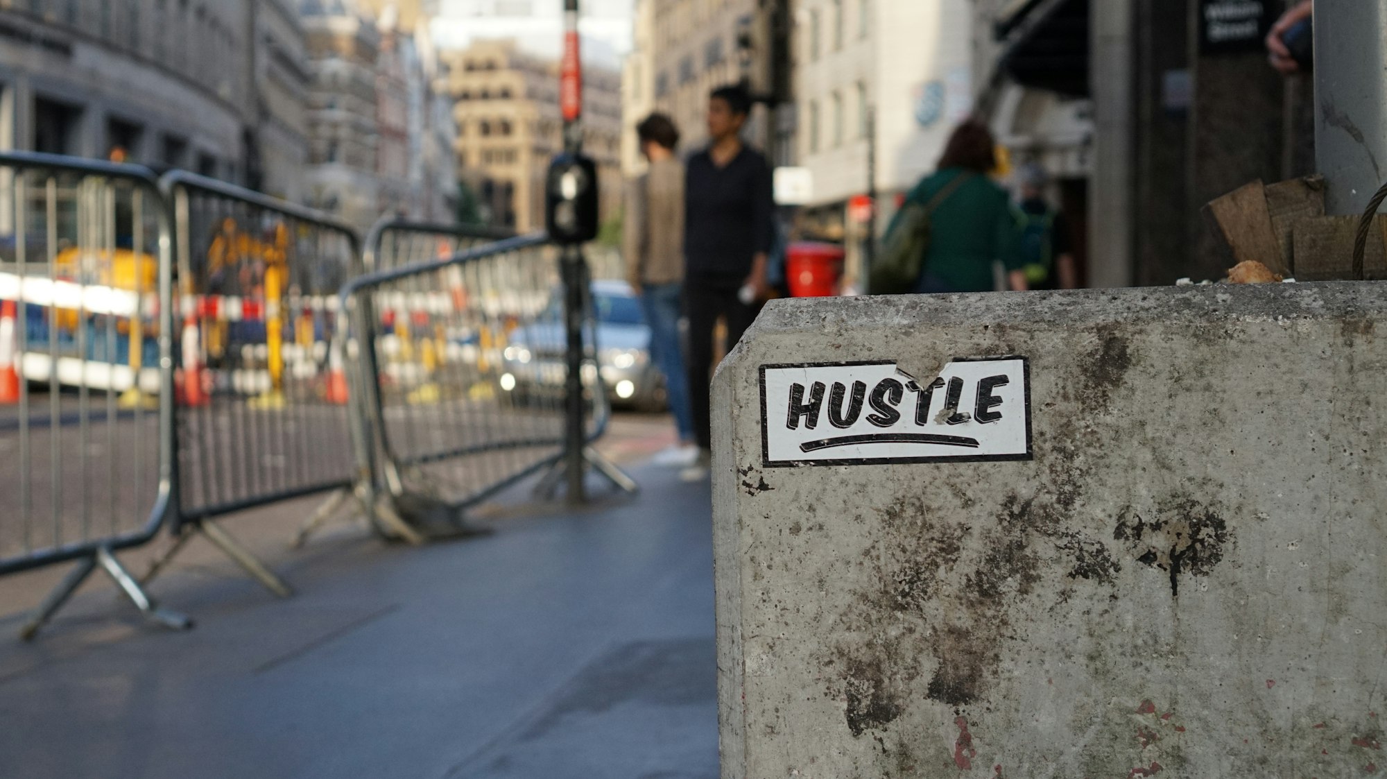 a sticker with the word "hustle" on a concrete block on a sidewalk, with barricades blocking lining the curb