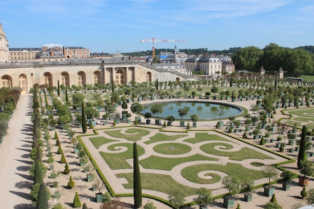 An ornate garden with precisely trimmed hedges and symmetrical designs surrounding a central reflective pond. The garden is bordered by a large historical building with arched windows, and several potted trees align the pathways. In the background, construction cranes and more architecture can be seen.
