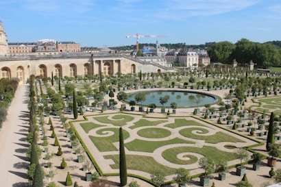 An ornate garden with precisely trimmed hedges and symmetrical designs surrounding a central reflective pond. The garden is bordered by a large historical building with arched windows, and several potted trees align the pathways. In the background, construction cranes and more architecture can be seen.