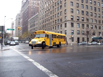 A yellow school bus turns a corner on a city street surrounded by tall brick buildings. The road is wet, indicating recent rain, and there are several cars and pedestrians in the vicinity.