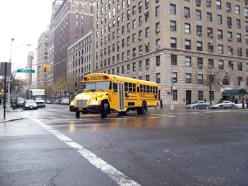 A yellow school bus turns a corner on a city street surrounded by tall brick buildings. The road is wet, indicating recent rain, and there are several cars and pedestrians in the vicinity.