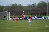 A women's soccer match is taking place with players on the field wearing red and blue uniforms. One player is leaping to head the ball near the goal with a goalkeeper and several other players nearby. The match is happening outdoors on a grass field, surrounded by a black metal fence and trees in the background.