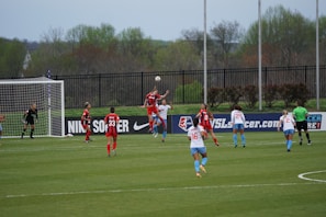 A women's soccer match is taking place with players on the field wearing red and blue uniforms. One player is leaping to head the ball near the goal with a goalkeeper and several other players nearby. The match is happening outdoors on a grass field, surrounded by a black metal fence and trees in the background.