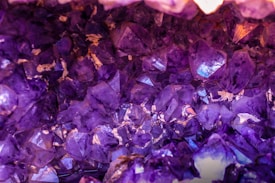 An array of purple crystals densely packed together, with light reflecting off their surfaces creating a vibrant and mysterious appearance. The crystals exhibit various shades of purple with some areas tinged by subtle hints of orange and white due to lighting and underlying minerals.