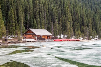 A wooden cabin situated at the edge of a semi-frozen lake with scattered ice, surrounded by dense evergreen forest. Nearby, a row of red canoes is stacked on a dock. A few people are visible near the cabin, likely preparing for an outdoor activity. The overall scene suggests a rustic, tranquil setting in a secluded area.