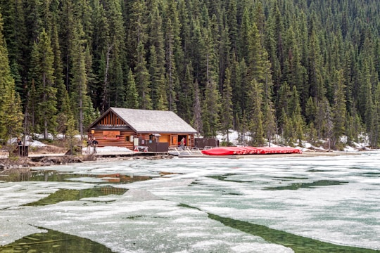 A wooden cabin situated at the edge of a semi-frozen lake with scattered ice, surrounded by dense evergreen forest. Nearby, a row of red canoes is stacked on a dock. A few people are visible near the cabin, likely preparing for an outdoor activity. The overall scene suggests a rustic, tranquil setting in a secluded area.