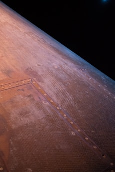 A close-up view of a large, textured surface with hexagonal patterns and rivets, likely part of a spacecraft or aircraft. The material appears metallic and has a worn, weathered appearance. The background is mostly dark, suggesting space or a high altitude.