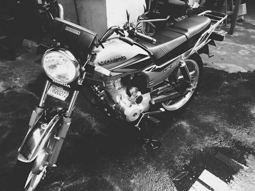 A motorcycle with a Honda logo is parked on a wet surface. The image is in black and white, highlighting the bike's design and chrome detailing. The vehicle appears clean and polished, reflecting the surroundings on its metallic parts.