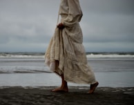 Models walking barefoot on the beach wearing flowing kaftans with ocean waves in the background.