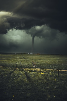 A tornado is prominently visible under a dark, stormy sky over an expansive green field. The clouds are heavy and ominous, creating a dramatic and intense atmosphere.