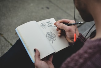 A person is sketching intricate designs in a notebook while seated outdoors. The hand holds a pencil, drawing detailed illustrations accompanied by handwritten text.