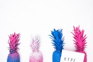 Four pineapples painted in bright and bold colors, including pink, blue, and a combination of blue and pink, are arranged against a white background. A white lightbox with the word 'HYPE' is placed in front of the pineapples, adding a playful and modern touch.
