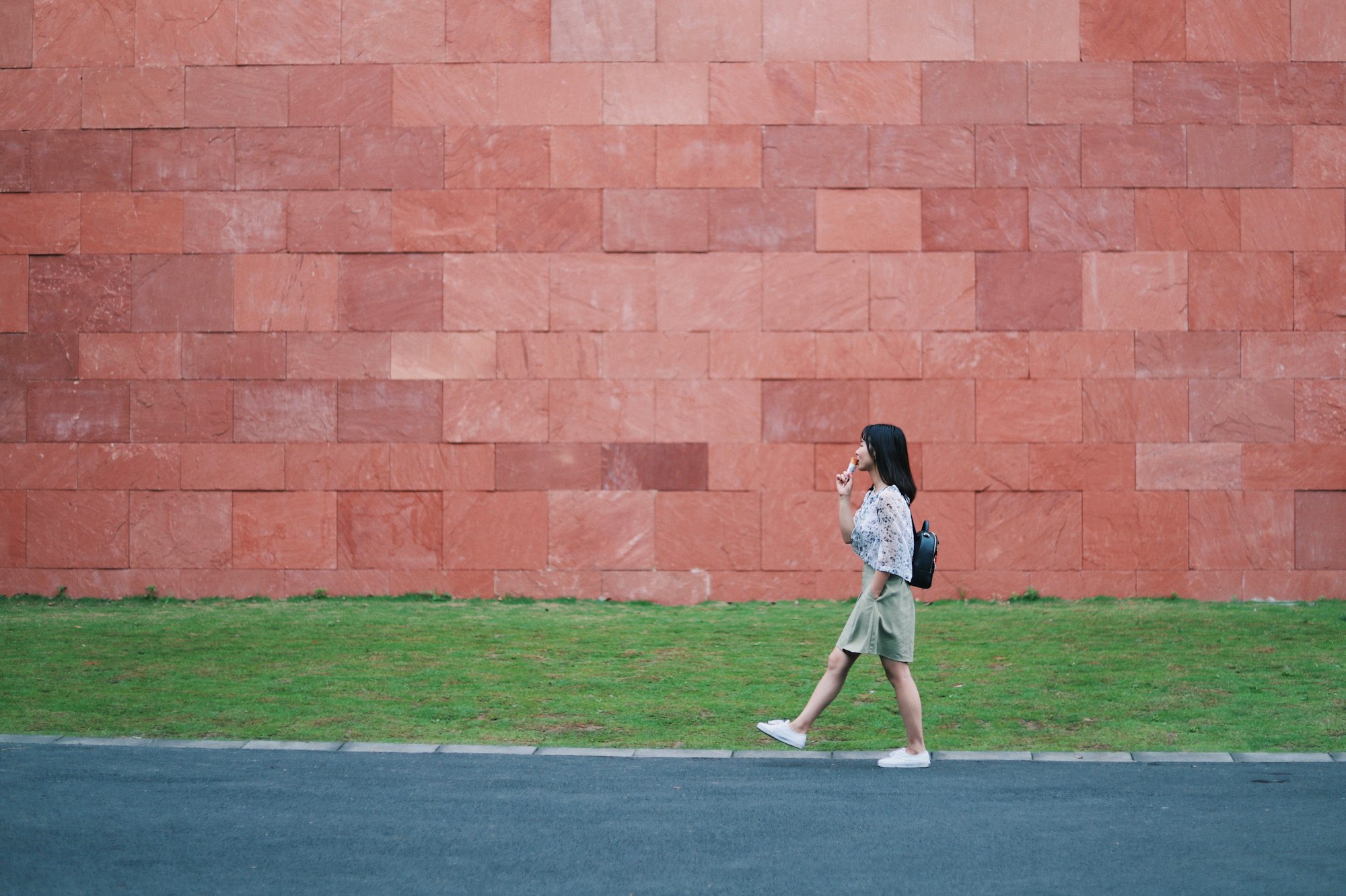 Jeune femme marche à pied et mange une glace devant un grand mur rouge