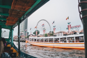 A lively riverside scene with a covered boat in the foreground navigating the water. On the opposite bank, a ferris wheel towers above a bustling area adorned with several flags. People are visible on the pier and on another boat. A large sign reading 'Asiatique' is displayed on top of a building. High-rise buildings loom in the background, indicating an urban setting.