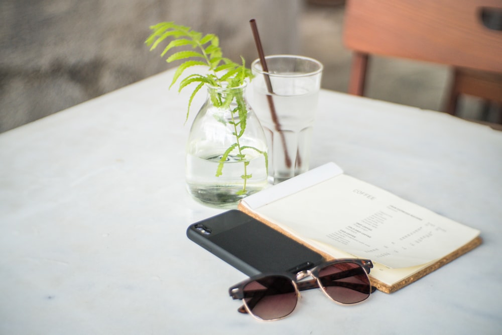 black framed sunglasses beside black cased smartphone and white covered book on white table